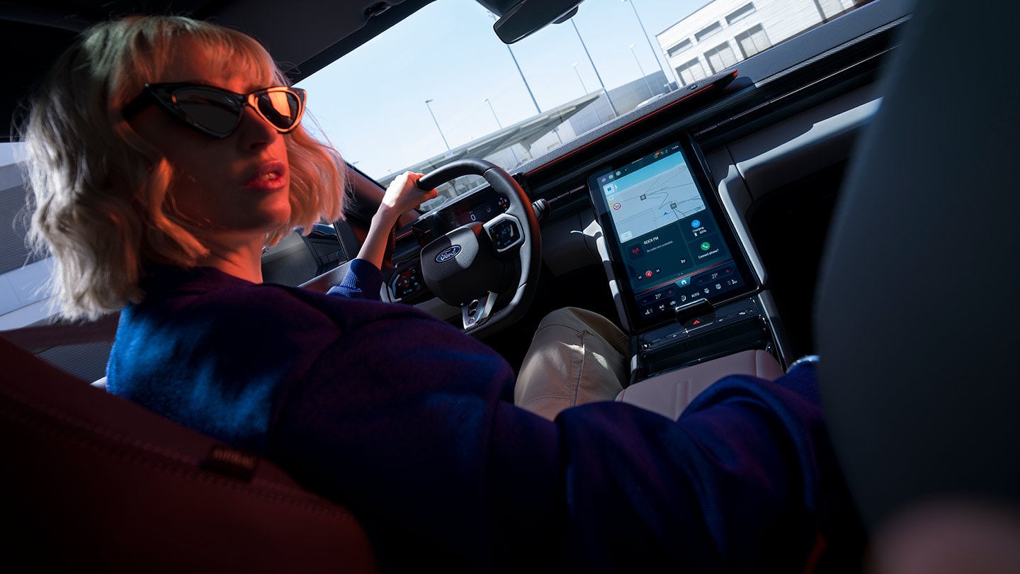 Eine Frau fährt den vollelektrischen Ford Capri®. Zu sehen sind das Lenkrad, das Armaturenbrett und der grosse Ford SYNC® Move-Touchscreen.