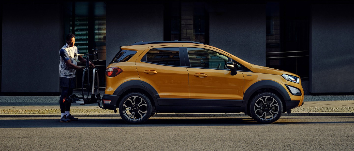 Ford EcoSport in orange Seitenansicht parkend auf Strasse in Stadt während ein Mann ein Fahrrad auf dem Heckträger befestigt.